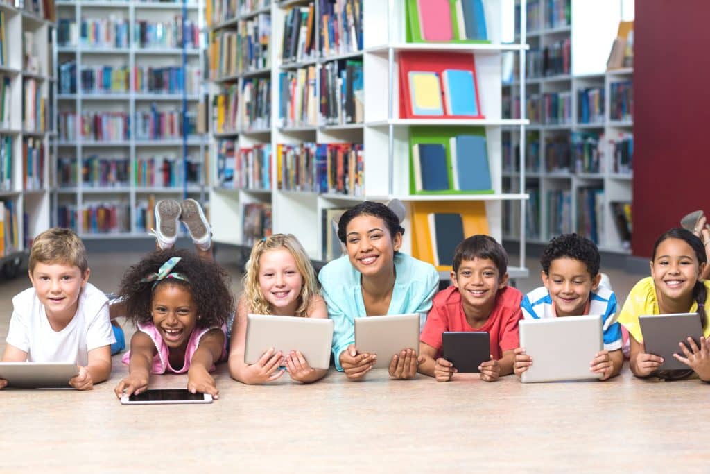Portrait of smiling teacher with students using digital tablets while lying down in library | Edsby Portrait of smiling teacher with students using digital tablets while lying down in library