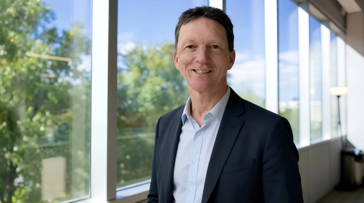 Smiling man in a dark suit and light blue shirt standing beside large office windows.